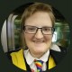 Clare O'Hanlon smiling white person with glasses, short light brown hair, rainbow tie, dark jacket with yellow collar 
