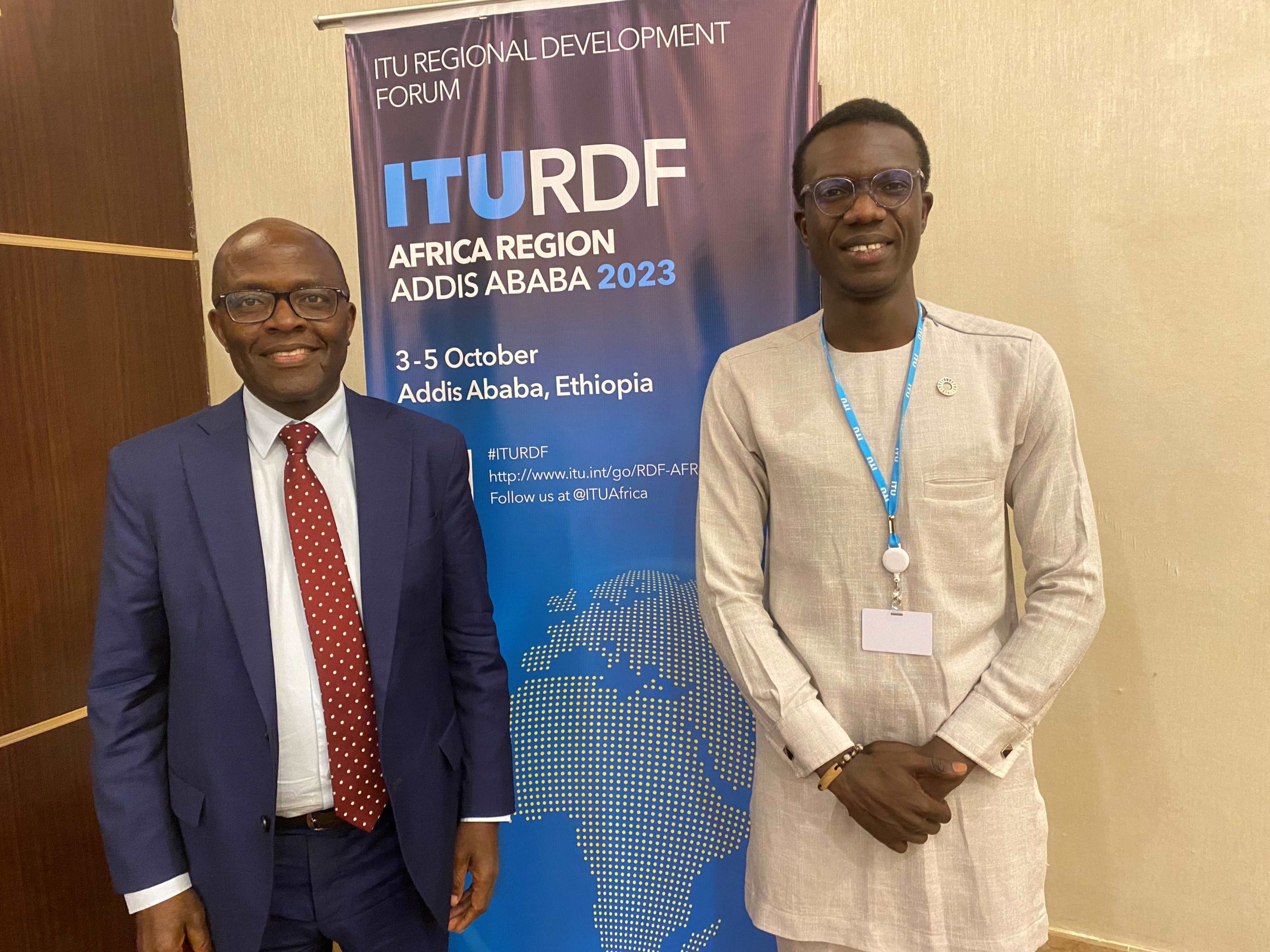 Two African men. On the left, a smiling man with a dark blue suit, white shirt and red spotted tie. On the right, another smiling man in a beige top. Behind them, a banner with ITURDF and an image of Africa